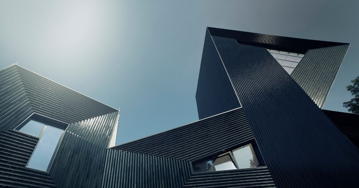 The image shows a modern, angular building with metallic surfaces, photographed from a low angle against a bright sky