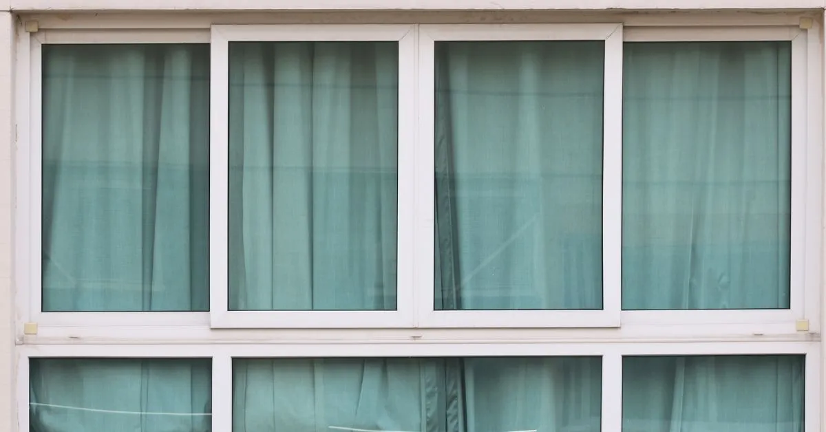 A white aluminium window with multiple glass panes, partially covered by light teal curtains, showcasing a simple and classic design.